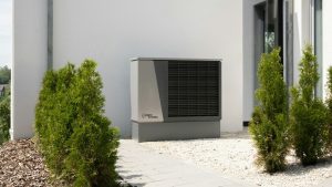 Heat pump installed against house wall with plants, tiles and stones closer to the foreground on a sunny day.