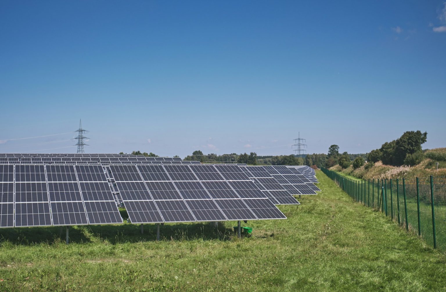 Solar panel arrays (one after another heading into the distance) in a field next to a green metal fence with a blue sky in the background as well as power cables in the distance.