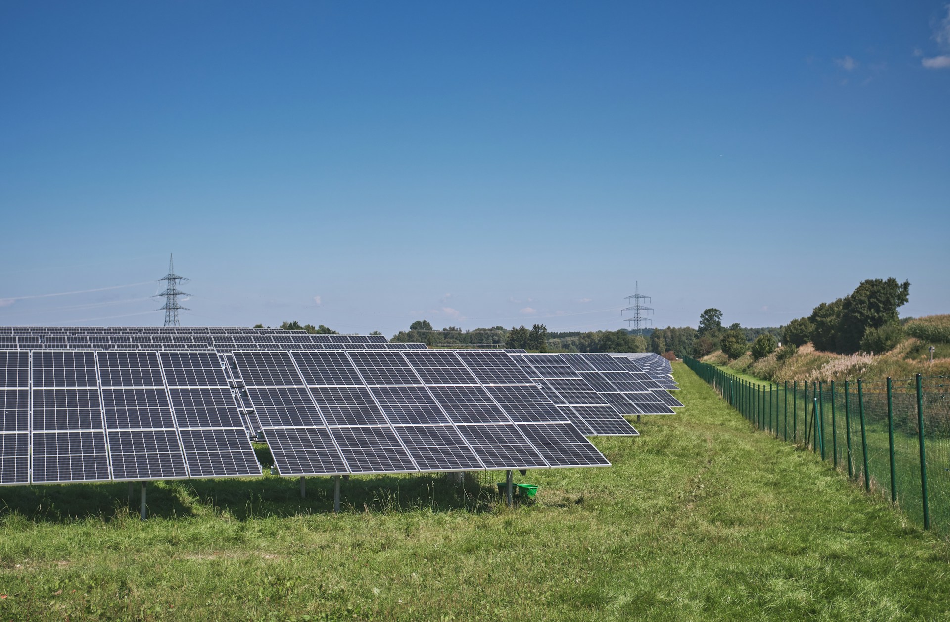 Solar panel arrays (one after another heading into the distance) in a field next to a green metal fence with a blue sky in the background as well as power cables in the distance.