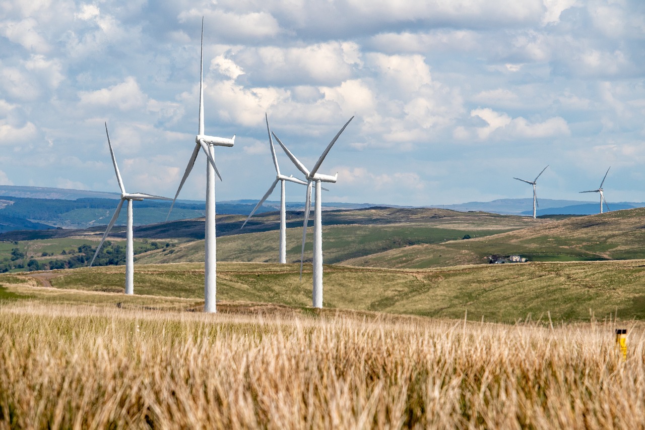 Wind turbines in a field on a day with some cloud cover but plenty of brightness too. Some hay in the foreground and hills in the background.